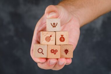 Pessoa segurando blocos de madeira empilhados com ícones representando pesquisa, processos, ideias, metas, dinheiro e sucesso, simbolizando o desenvolvimento de soft skills